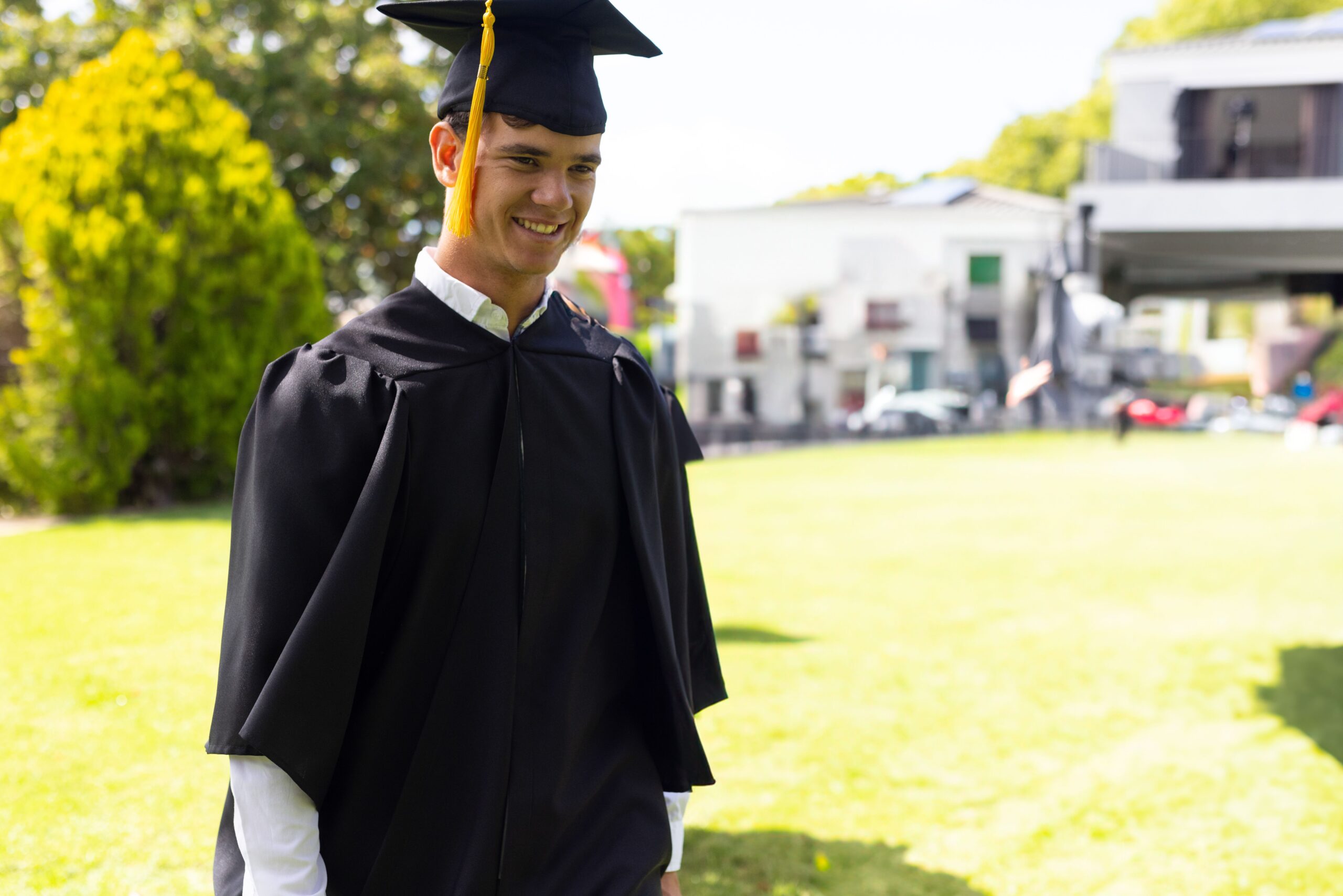 White male graduate wearing black gown with mortarboard cap walking across campus lawn, copy space